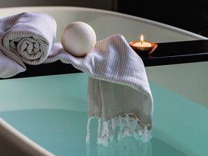 A semi-unrolled towel, bath bomb, and lit candle is perched on a piece of wood that stretches over a calm filled spa.